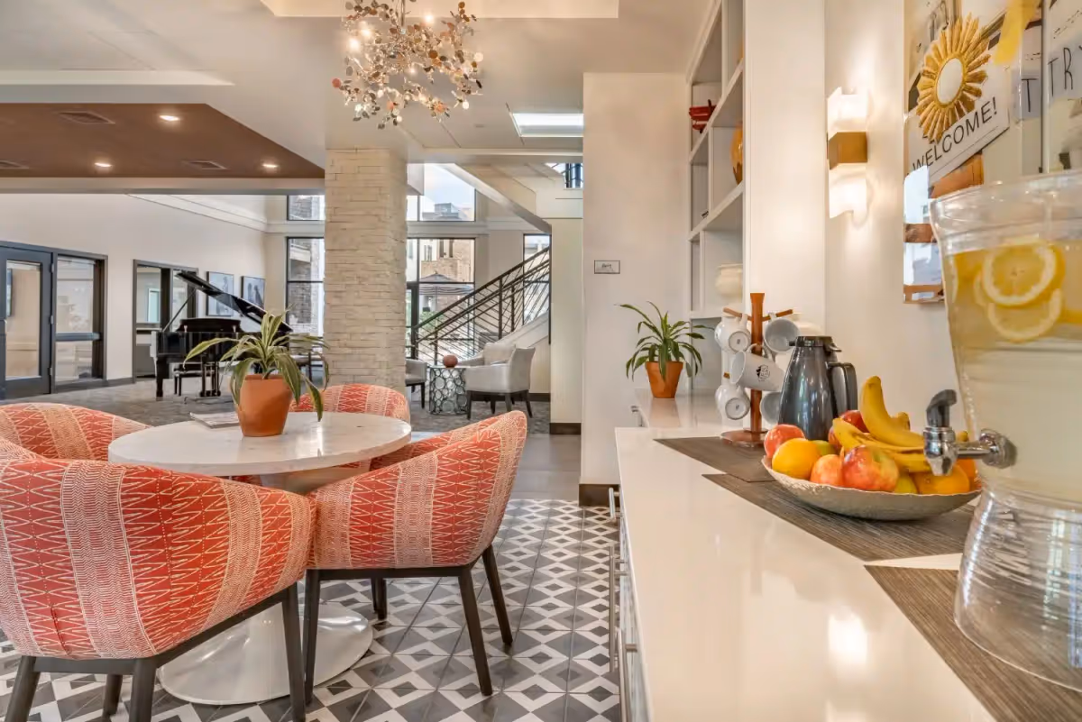 A bright and welcoming common area in a senior living facility featuring a round table with four patterned red chairs, a potted plant centerpiece, a side counter with a bowl of fresh fruit, a coffee pot, mugs on a rack, and a large glass dispenser filled with lemon water. In the background, there is a grand piano, large windows, a stone pillar, and a seating area with armchairs near a staircase.