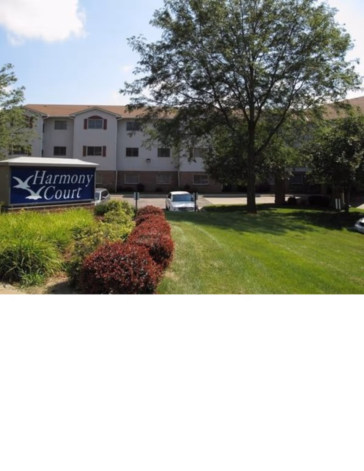 Front exterior of the Harmony Court senior living building with its sign, lawn, shrubs, and a large tree.