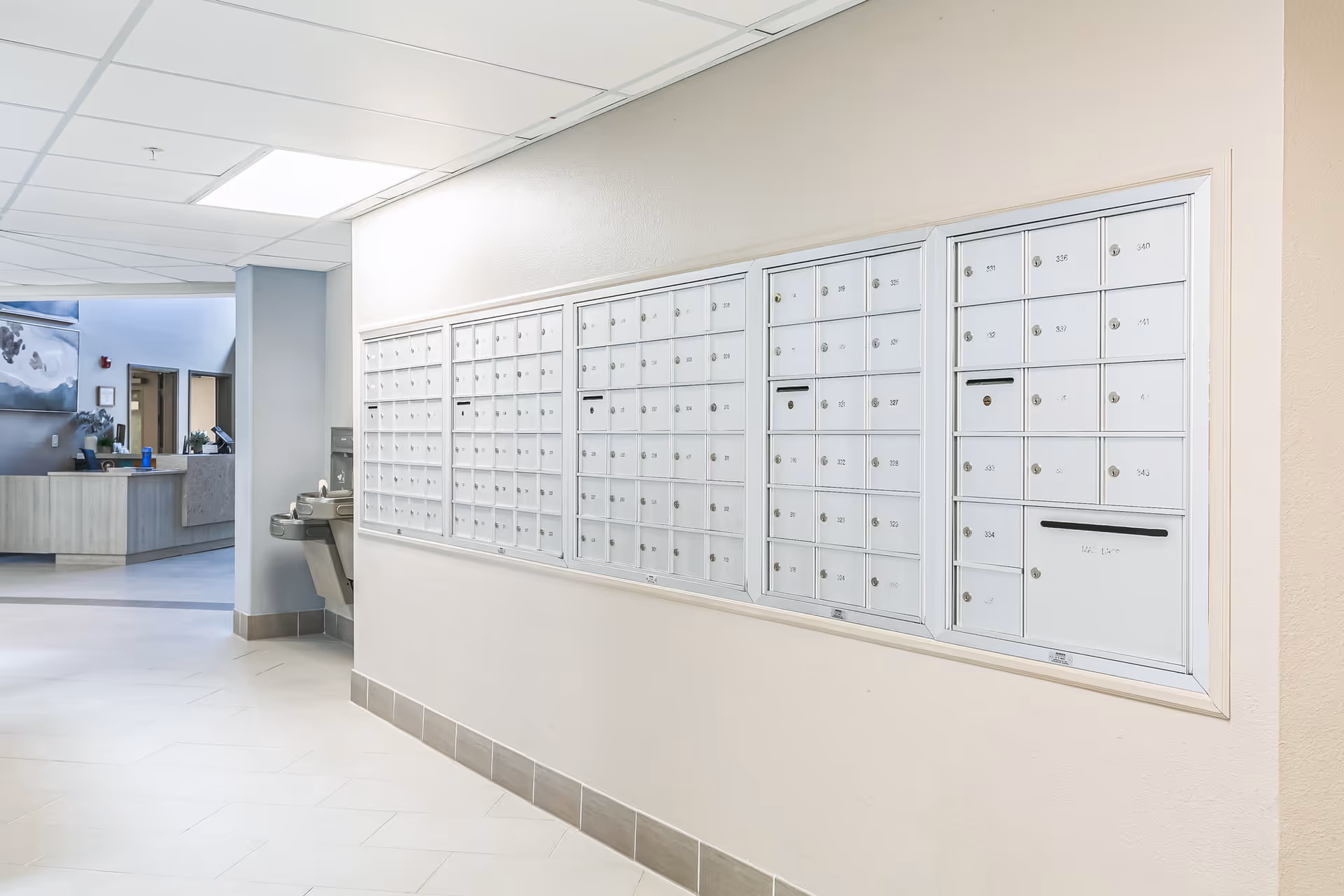 Interior hallway of a senior living facility featuring a wall-mounted bank of white mailboxes with numbered compartments. To the left, there is a water fountain and a reception desk area with a computer and some decor. The space is well-lit with white walls and tiled floors.