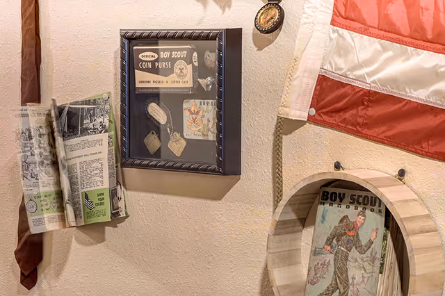 A wall display featuring Boy Scout memorabilia including a framed collection of Boy Scout coin purses, a hanging Boy Scout handbook, a folded newspaper clipping, a red and white flag, and a small compass.