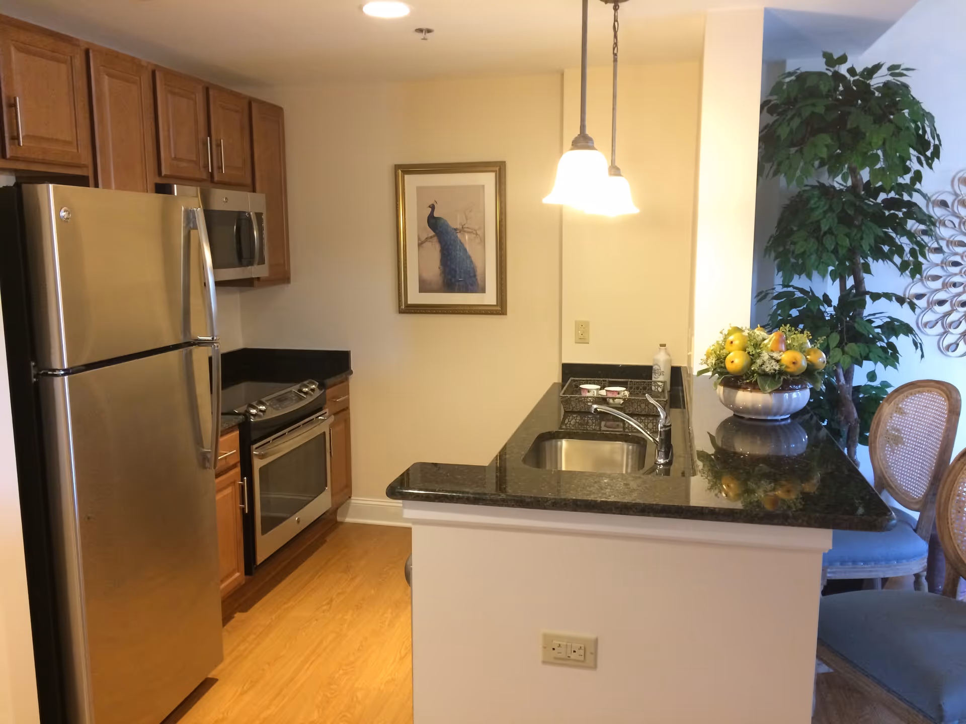 A modern kitchen with wooden cabinets, stainless steel refrigerator, oven, and microwave. There is a black granite countertop with a sink and a decorative fruit bowl. Two pendant lights hang above the counter, and a framed picture of a peacock is on the wall. Part of a dining area with chairs and a large green plant is visible in the background.