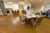 Interior view of a senior living facility common area with wooden flooring, several tables and chairs arranged for dining or social activities, a kitchen area visible in the background, and a pool table on the right side.