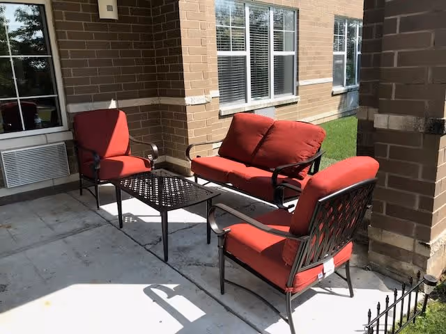 Outdoor patio area with red cushioned metal chairs and a loveseat arranged around a black metal table, adjacent to a brick building with windows and a grassy area nearby.