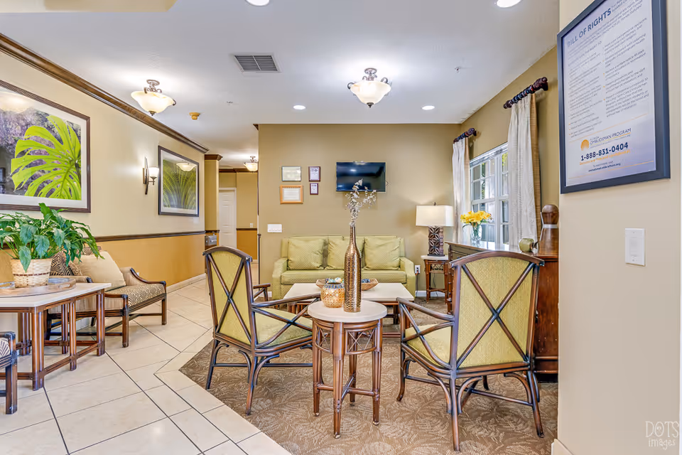A cozy living room area in a senior living facility with a green sofa, two green cushioned chairs, a coffee table, and side tables. The room has beige walls with framed artwork and a window with curtains letting in natural light. There are ceiling lights and a wall-mounted TV above the sofa.