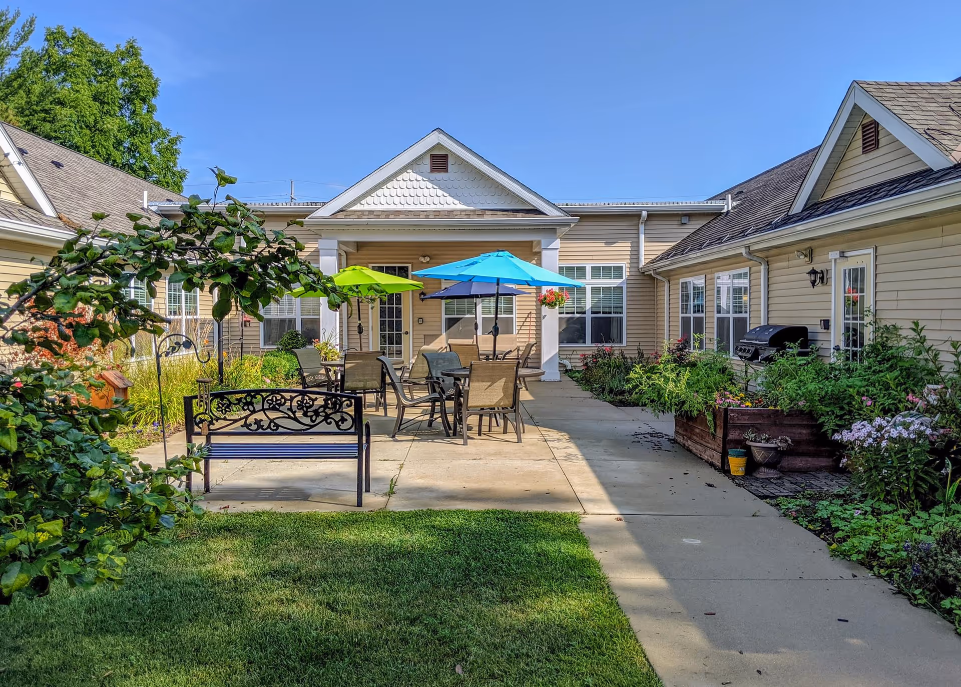 Outdoor patio area at a senior living facility with a bench, several tables and chairs under colorful umbrellas, surrounded by garden beds and greenery, with beige buildings in the background under a clear blue sky.