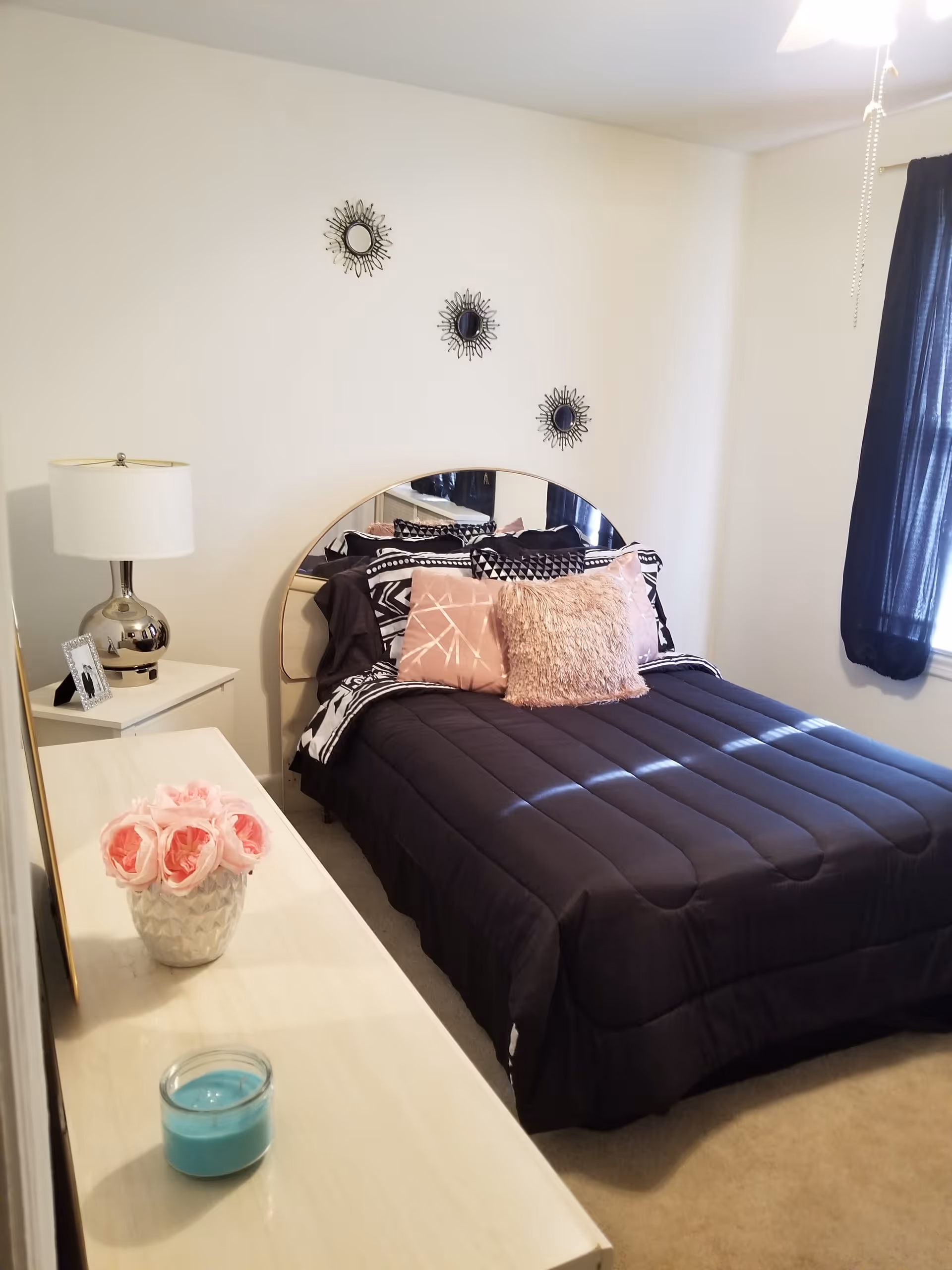 A cozy bedroom with a neatly made bed featuring black bedding and decorative pillows in pink and black patterns. Above the bed are three small sunburst mirrors arranged diagonally on a white wall. To the left of the bed is a white nightstand with a silver lamp and a framed photo. In the foreground, there is a white dresser with a vase of pink roses and a blue candle. A window with dark curtains is on the right side of the room.