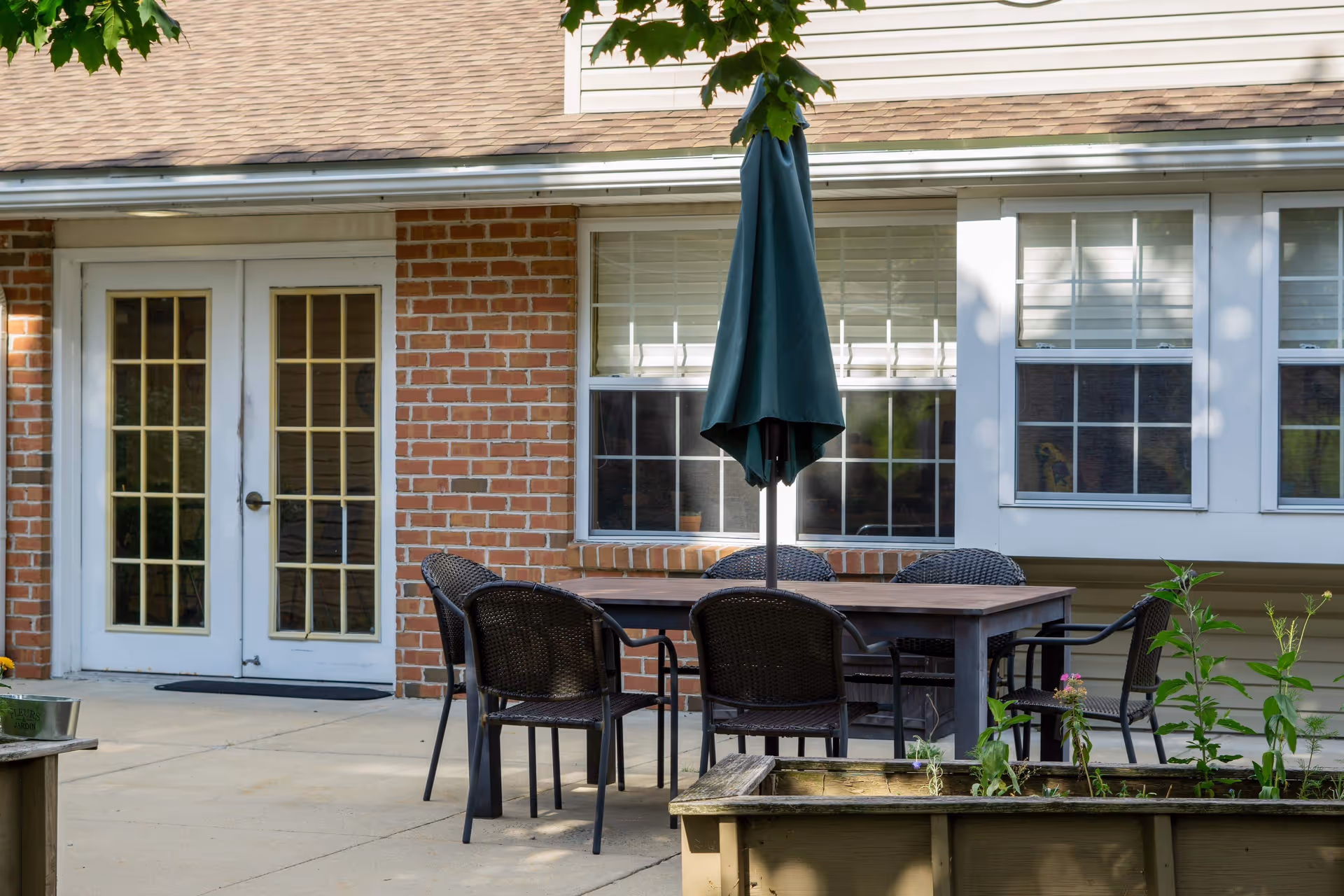 Outdoor patio area with a rectangular table surrounded by six wicker chairs and a closed green umbrella in the center. The patio is adjacent to a brick building with white-framed French doors and windows with white blinds. There is a raised garden bed with plants in the foreground.