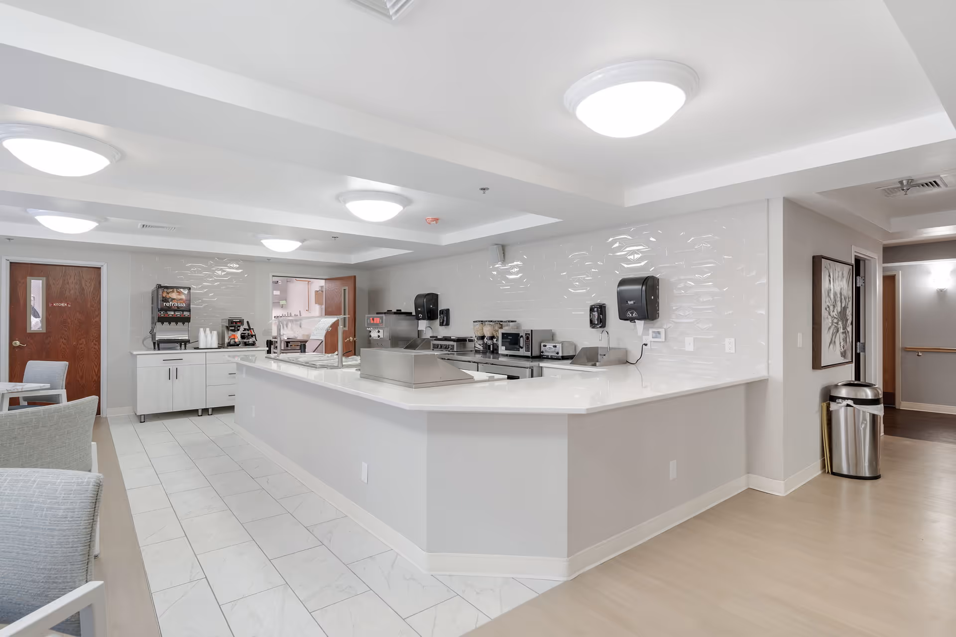 A clean, modern kitchen area in a senior living facility with white countertops, stainless steel appliances, and a beverage dispenser. There are light gray walls, tiled floors near the kitchen, and light wood flooring in the adjacent area. Chairs and tables are partially visible on the left side.
