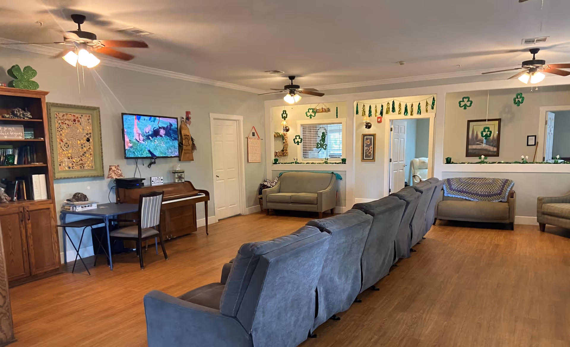A spacious living room with wooden flooring, multiple gray recliner chairs arranged in a row, two ceiling fans with lights, a wall-mounted TV showing a nature scene, a piano, a small table with a chair, and green shamrock decorations hanging from the ceiling and walls.