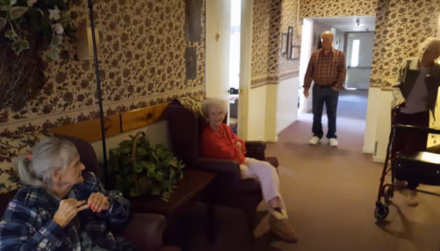 Several elderly residents sit and stand in a carpeted common area/hallway with floral wallpaper.