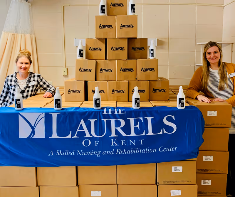 Two women standing behind a table covered with a blue banner that reads 'The Laurels of Kent A Skilled Nursing and Rehabilitation Center.' Behind them is a pyramid stack of cardboard boxes labeled 'Amway' with white bottles placed on top and in front of the boxes.