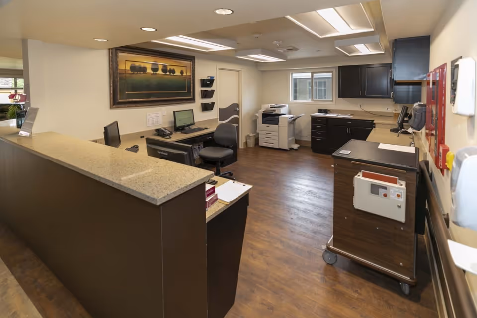 Interior view of a reception or nurse station area in a senior living facility with a long counter, office chairs, computer monitors, a printer, cabinets, and a cart on wheels. The room has wood flooring, beige walls, and ceiling lights. A framed landscape painting hangs on the wall.