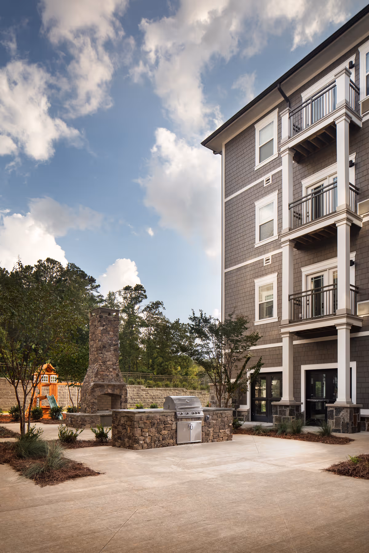 Outdoor patio area at Monark Grove Greystone featuring a stone fireplace, a built-in stainless steel grill, trees, and a multi-story building with balconies under a partly cloudy sky.