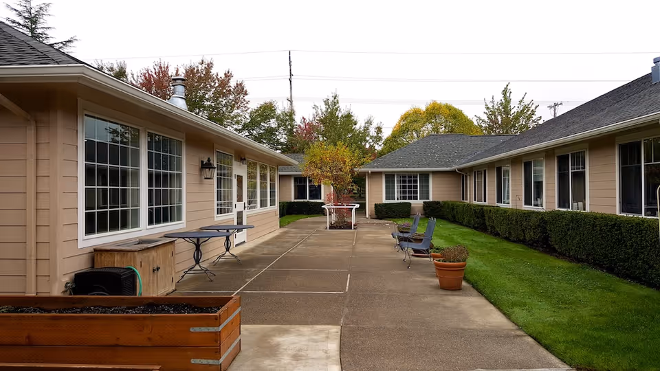 Outdoor courtyard area between two beige single-story buildings with large windows. The courtyard has a concrete walkway, several chairs, potted plants, a small tree in the center, and a raised garden bed. Green grass and trimmed hedges line the walkway, with trees visible in the background under an overcast sky.