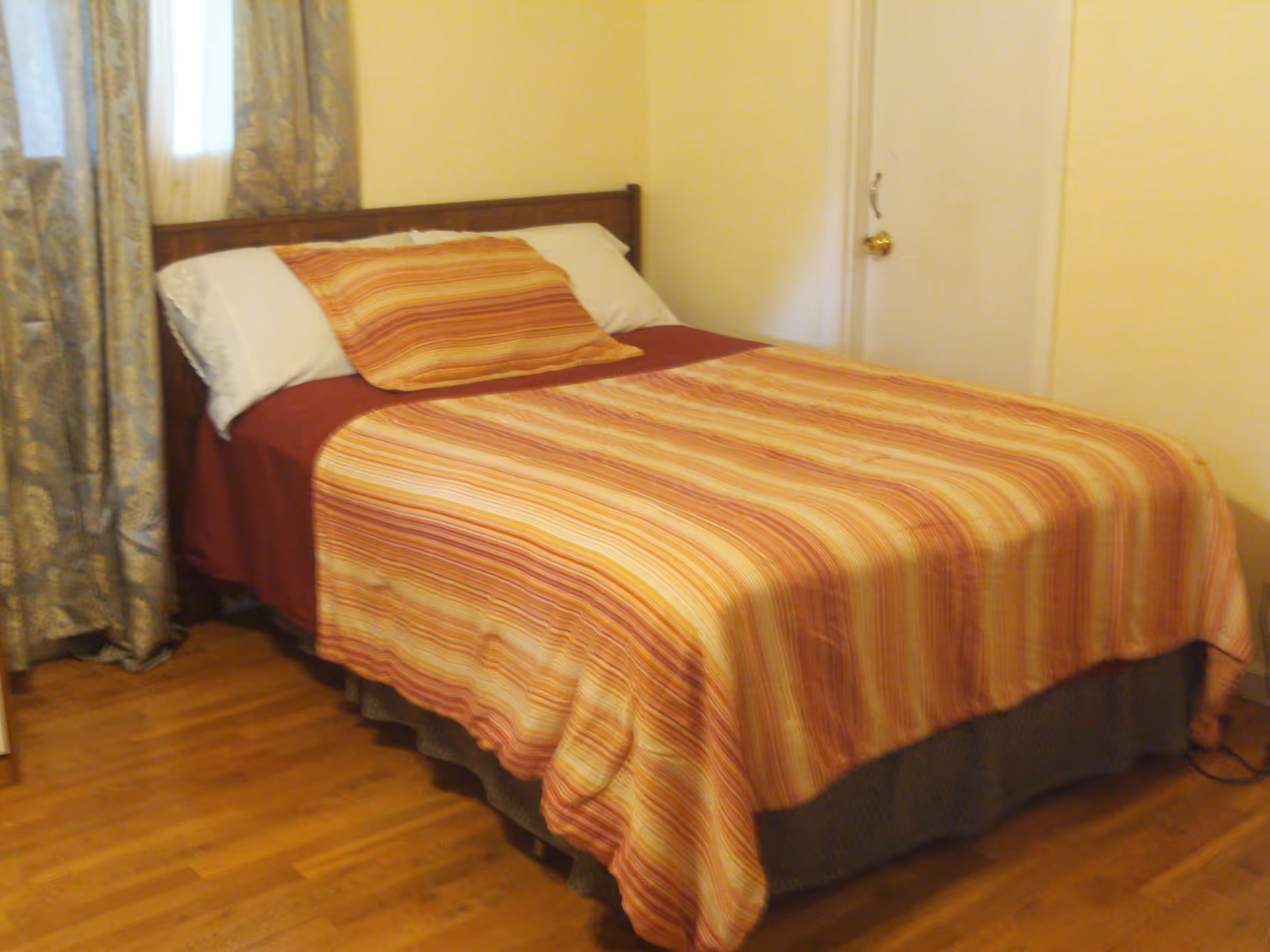 A bedroom with a wooden bed frame, a bed made with a striped orange, yellow, and red bedspread, and matching pillow. The room has light-colored walls, a wooden floor, a window with patterned curtains, and a closed white door with a brass doorknob.