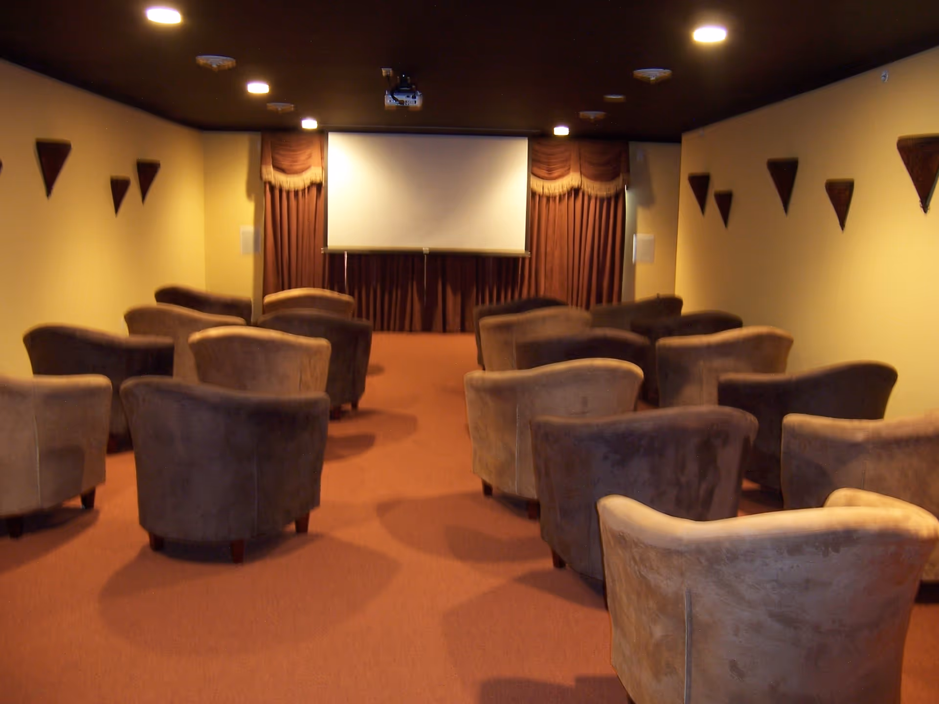 A small theater room with multiple rows of brown upholstered chairs facing a large white projection screen with brown curtains on the wall behind it. The room has beige walls with decorative triangular wall hangings and recessed ceiling lights.