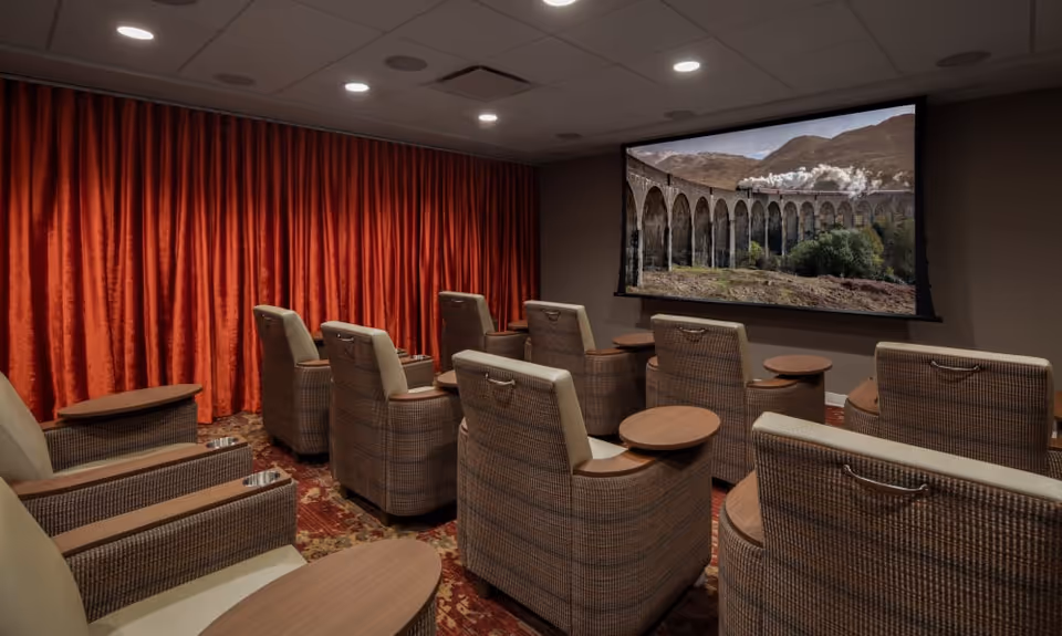 A small theater room with two rows of cushioned armchairs facing a large screen displaying a scenic image of a steam train crossing a stone viaduct. The room has a patterned carpet, red curtains along one wall, and recessed ceiling lights.