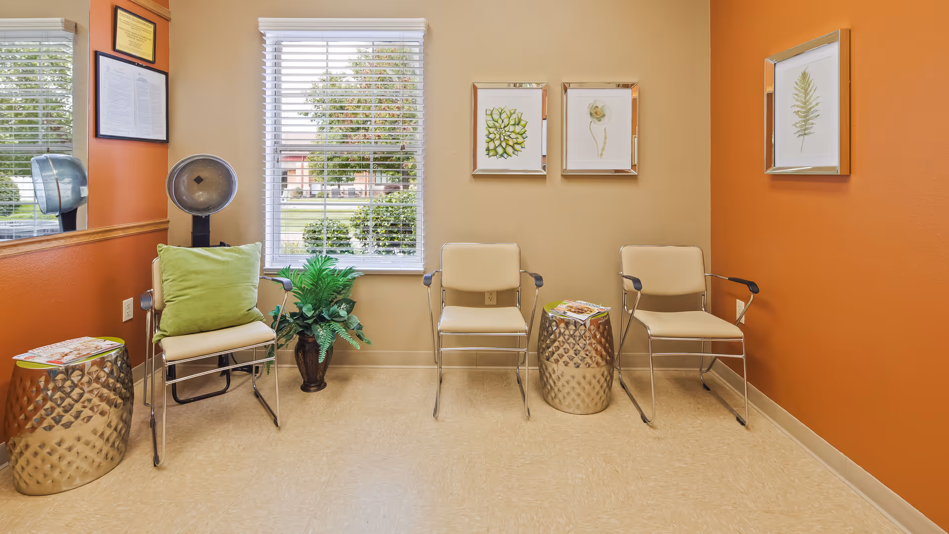 A small waiting area with three beige chairs, two metallic side tables with magazines, a green pillow on one chair, a potted plant, and framed botanical prints on the walls. There is a window with blinds showing greenery outside, and the walls are painted in warm beige and orange tones.