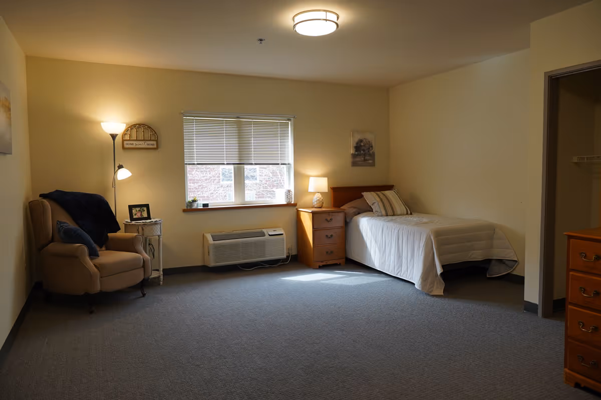 A simple, cozy bedroom in a senior living facility with a single bed covered in a white quilt, a wooden nightstand with a lamp, a window with blinds, a beige armchair with a dark blue blanket and pillow, a floor lamp, and a small side table with a framed photo. The room has beige walls and gray carpeted floor.