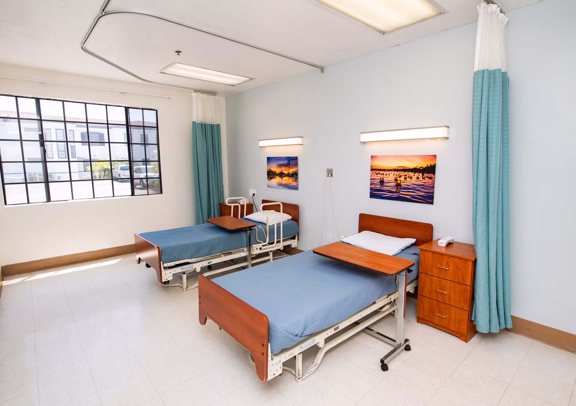 Two hospital-style beds with blue bedding, wooden headboards and overbed tables in a bright senior care patient room with teal privacy curtains and a large window.