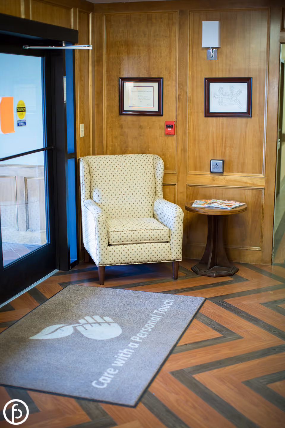 Wood-paneled lobby corner with an upholstered armchair, small round side table with magazines, and a welcome mat reading "Care with a Personal Touch".