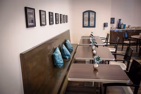 Interior view of a dining area with wooden tables and chairs. A long wooden bench with blue decorative pillows is positioned against a wall adorned with framed pictures. Small vases with blue flowers are placed on each table. In the background, there is a counter with decorative items and a wall-mounted phone.