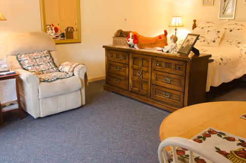 A cozy room with a beige armchair with a floral cushion and a folded blanket, a wooden dresser with drawers topped with a lamp, a stuffed dog toy, and framed photos. A bed with a white quilt and pillows is partially visible. A round table with a floral placemat and a white chair are in the foreground.