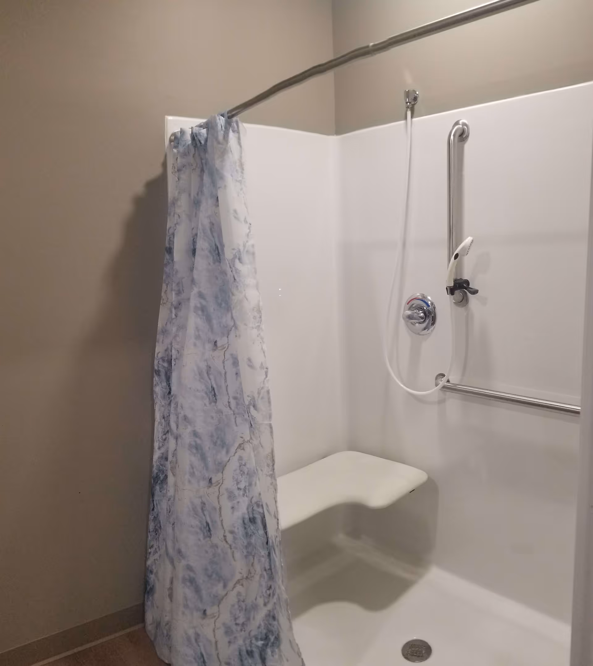 Accessible shower stall with built-in bench, handheld showerhead, grab bars, and a patterned shower curtain.