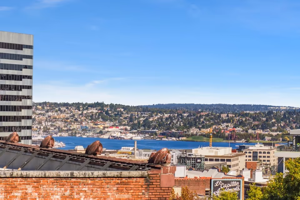 View over city rooftops toward a waterfront and hillside neighborhood under a clear blue sky.
