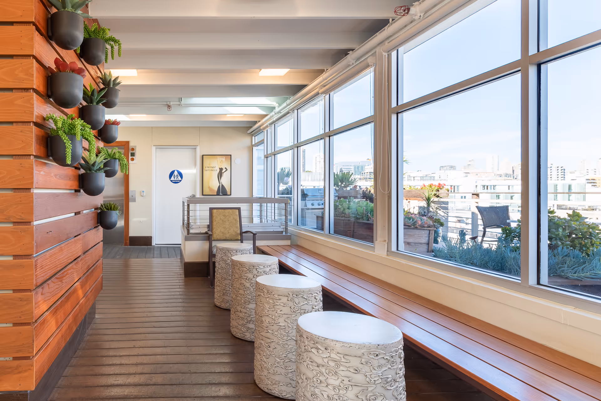 A bright indoor seating area with large windows overlooking an outdoor patio with plants and city buildings in the background. The room features a wooden bench along the window, four decorative white cylindrical stools, a single armchair, and a wall with multiple small potted plants. A restroom door and elevator are visible at the far end of the room.