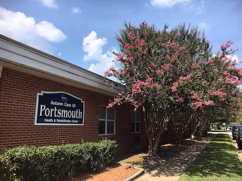 Exterior view of Autumn Care of Portsmouth Health & Rehabilitation Center, a single-story brick building with a sign on the wall. There are flowering trees with pink blossoms and a sidewalk leading alongside the building under a partly cloudy blue sky.