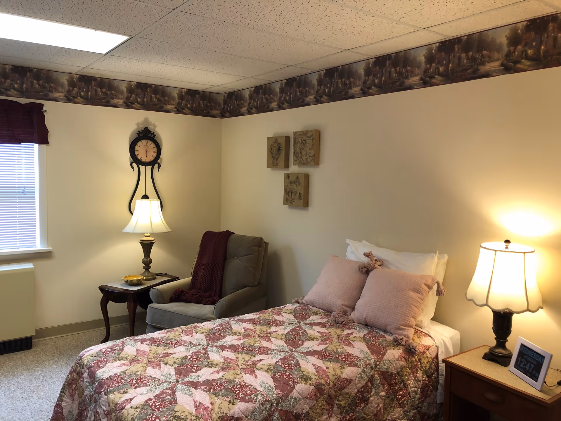 A cozy bedroom with a bed covered in a patterned quilt and two pink pillows. There is a cushioned armchair with a maroon throw blanket next to a small wooden side table with a lamp and a decorative bowl. On the wall above the chair is a decorative clock and three small framed artworks. Another lamp sits on a wooden nightstand beside the bed, which also holds a digital clock displaying the time 12:47. A window with blinds and a maroon valance is on the left side of the room.