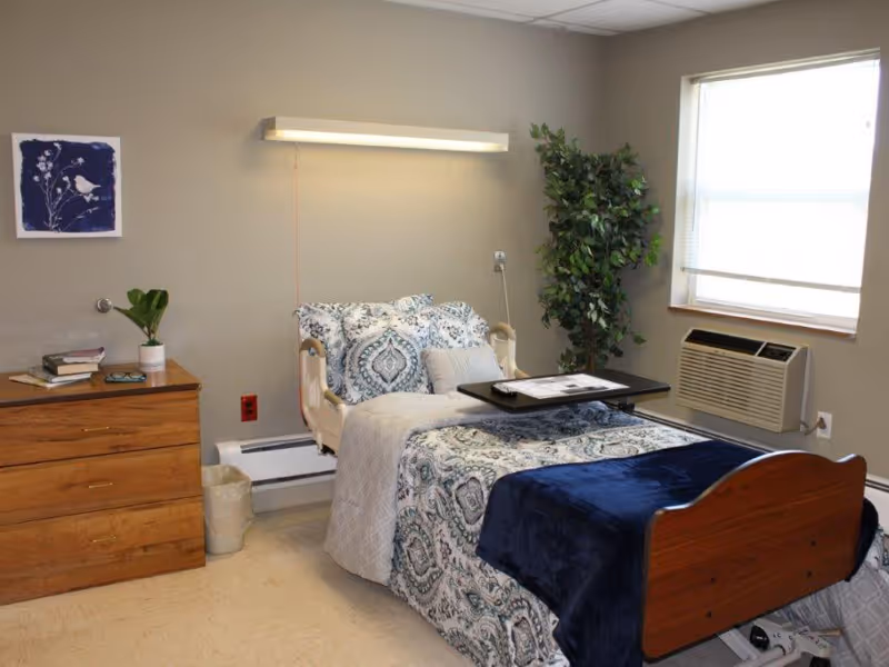 A neatly made single nursing facility bed with patterned bedding, overbed table, dresser, window air conditioner and a potted plant.