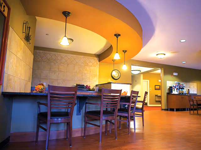 Interior view of a senior living facility reception or common area with a curved counter and four wooden chairs with dark cushions. The ceiling above the counter has a curved yellow design with three hanging pendant lights. The walls are tiled and painted in warm tones, and there is a clock on the wall behind the counter. In the background, there is a hallway and a small area with a coffee or beverage station and additional seating.