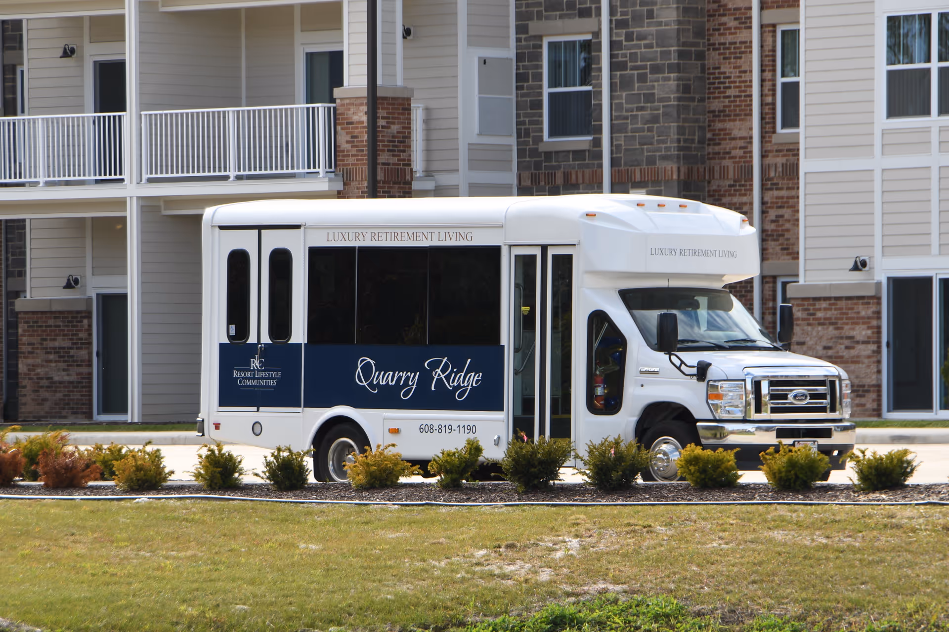 A white shuttle bus with blue signage reading 'Quarry Ridge' and 'Luxury Retirement Living' parked in front of a multi-story residential building with balconies and windows. There are small bushes and a grassy area in the foreground.