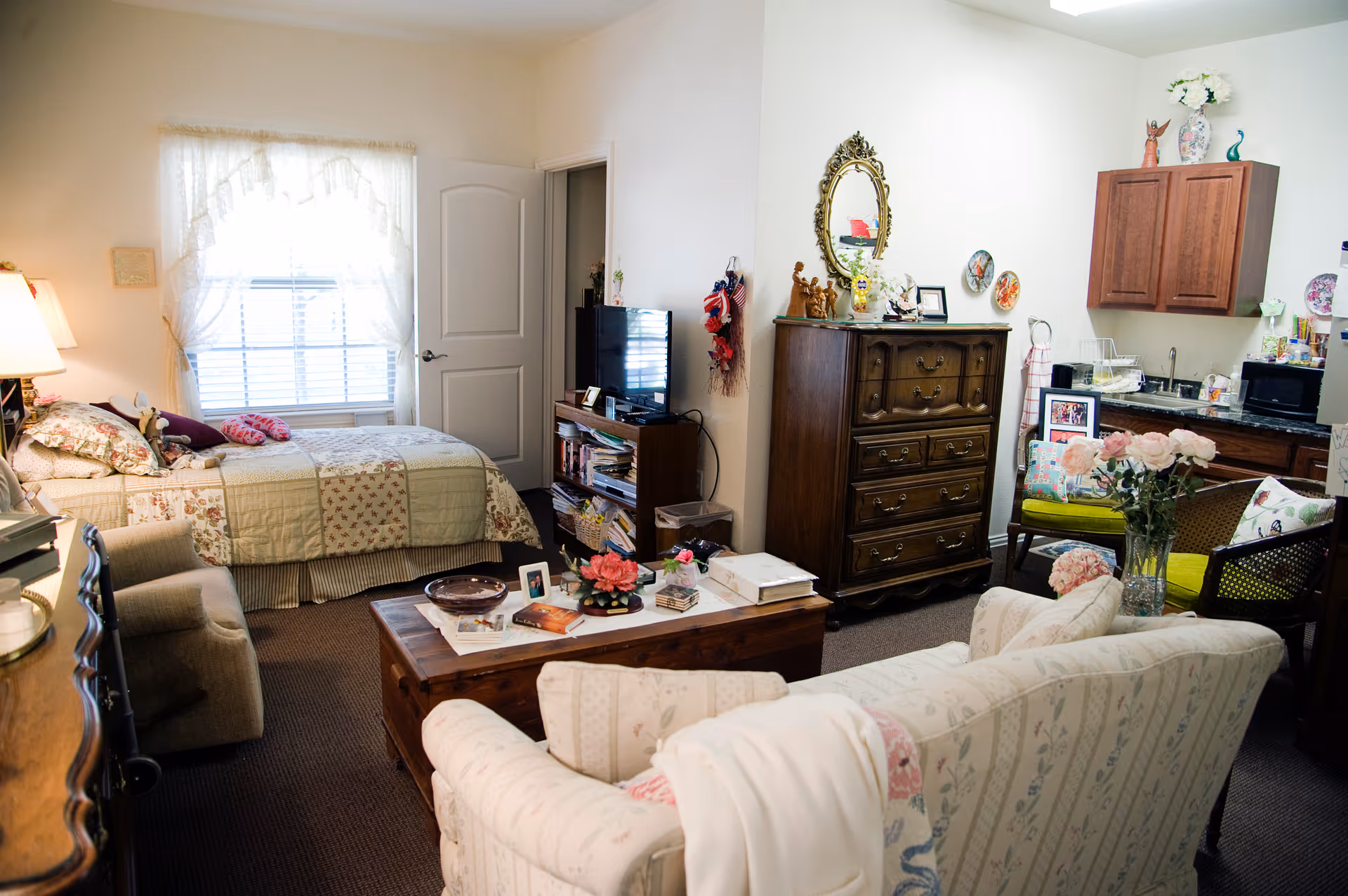 A cozy assisted living room with a bed near a window covered with lace curtains, a wooden dresser with a mirror above it, a small TV on a stand, a coffee table with books and flowers, and comfortable armchairs and a sofa. The room also includes a small kitchenette area with a sink, microwave, and cabinets.