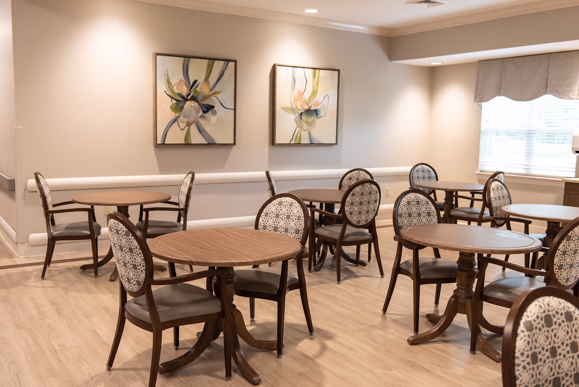A well-lit dining area with multiple round wooden tables and chairs featuring patterned backs and cushioned seats. The room has light-colored walls adorned with two floral paintings and a window with a valance allowing natural light to enter.