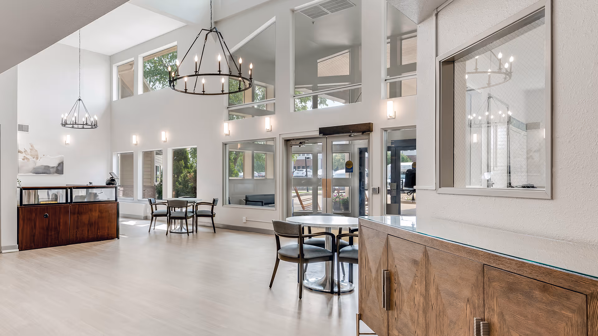 Bright and spacious interior of a senior living facility lobby with high ceilings, large windows, modern chandeliers, and seating areas with tables and chairs. The entrance doors and reception desk are visible, along with wooden cabinetry and light-colored flooring.