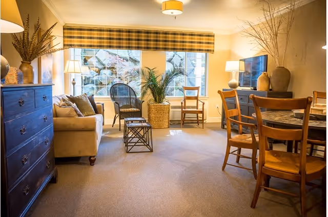 A cozy living room area with a beige sofa, multiple wooden chairs, a dark wooden dresser, a TV on a stand, and a round dining table with chairs. The room is warmly lit with lamps and natural light coming through large windows with a plaid valance. There are decorative plants and vases placed around the room.