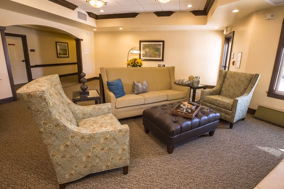 A cozy seating area in a senior living facility with a beige sofa adorned with two pillows, two patterned armchairs, a dark brown tufted ottoman with a tray on top, and side tables with decorative items. The room has beige walls, carpeted floor, and a framed picture on the wall.
