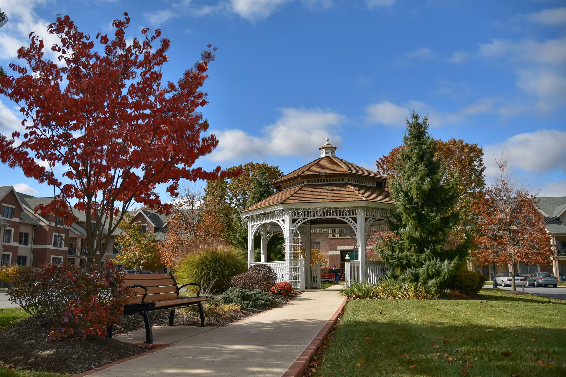 Outdoor scene at Phoebe Berks Health Care Center featuring a white wooden gazebo with a brown shingled roof, surrounded by autumn trees with red and orange leaves, green shrubs, and a bench along a paved walkway under a blue sky with some clouds.