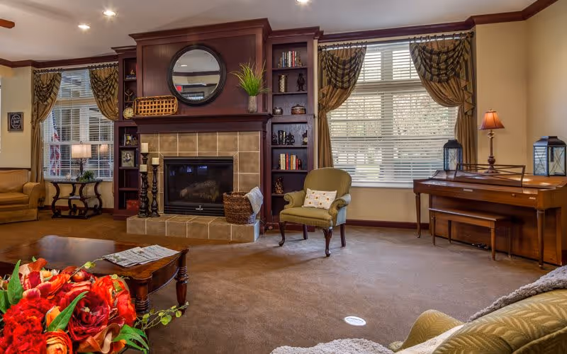 Cozy communal living room with a central fireplace, built-in shelving, seating, a piano, and large windows with drapes.