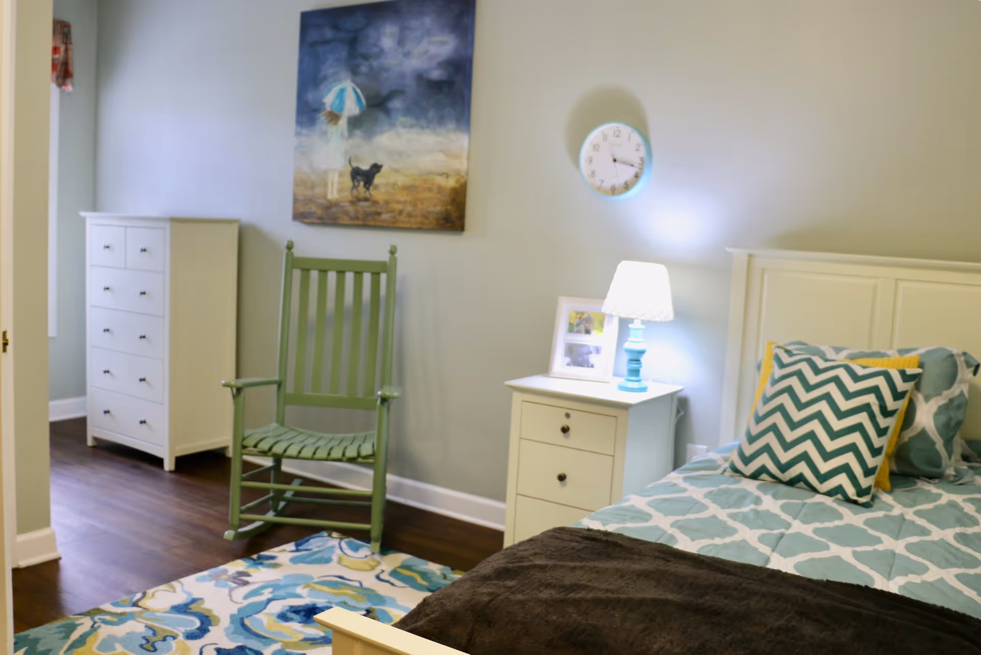 Bedroom with a single bed, patterned pillows and bedding, a bedside table with lamp, a green rocking chair, white dresser and wall art.