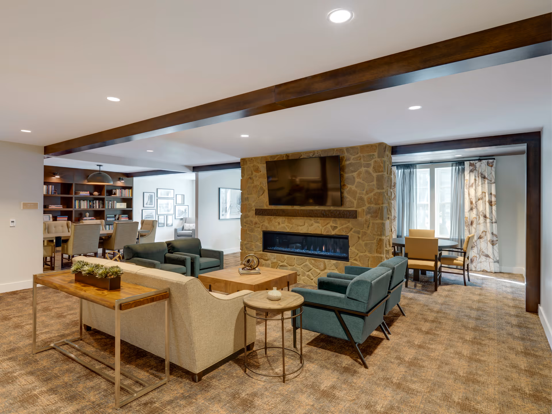 A cozy living room area featuring a stone fireplace with a mounted flat-screen TV above it. The room has a beige sofa, two teal armchairs, a wooden coffee table, and a side table. In the background, there is a dining area with a table and chairs near large windows with curtains, and a bookshelf with additional seating. The space has warm lighting and a carpeted floor.