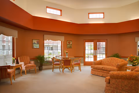 A spacious living room with orange walls and beige carpet. The room features a patterned orange sofa, matching armchairs, a wooden table with four chairs, and several potted plants. Large windows and glass doors let in natural light, and there are two small rectangular windows near the ceiling.