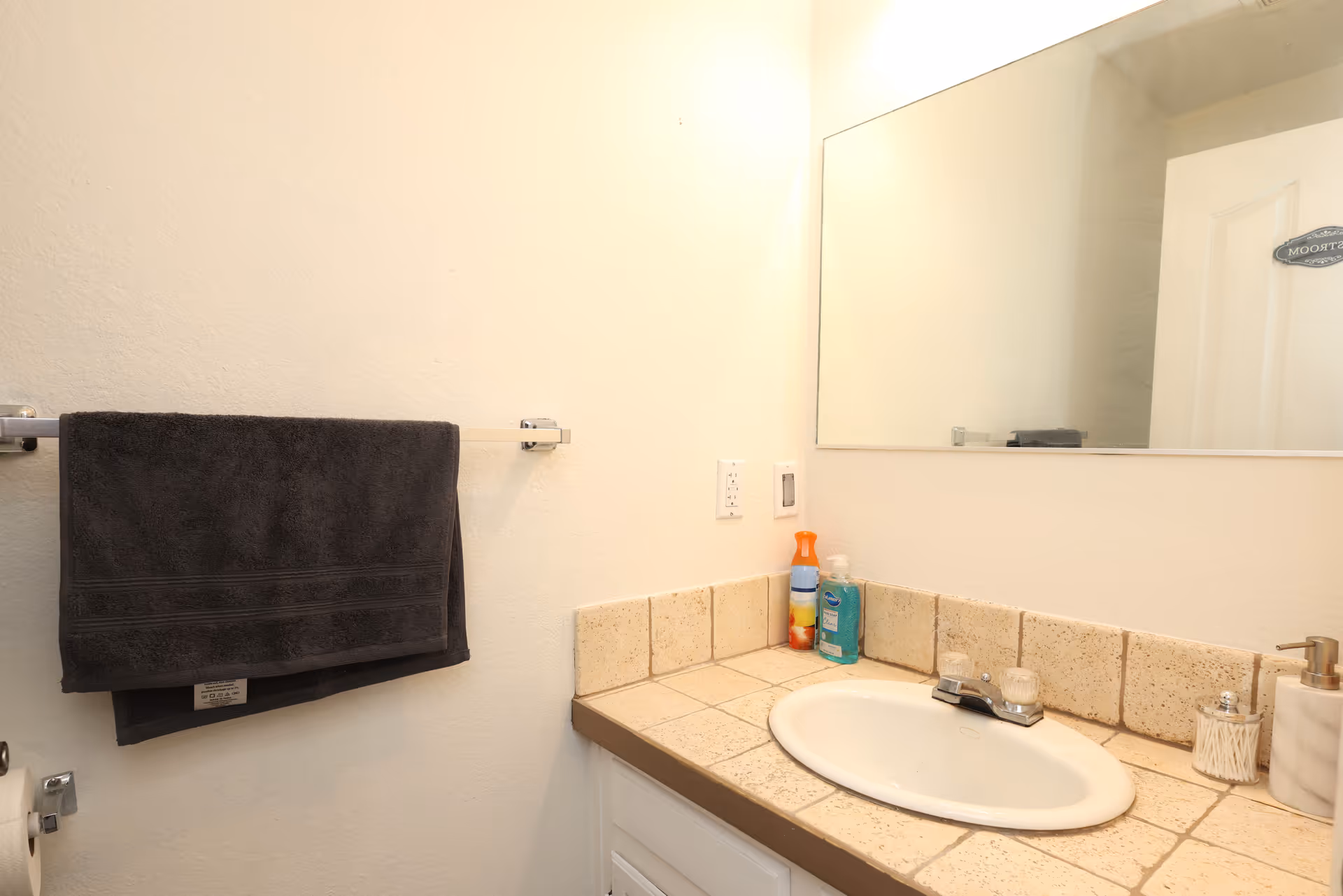 A small bathroom vanity with a tiled countertop, oval sink, mirror, and a dark towel hanging on a rack.