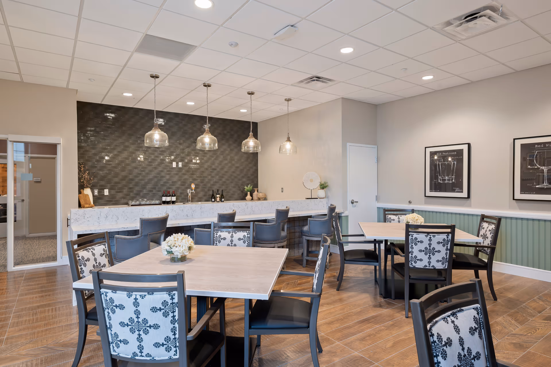 Bright communal dining room with tables and patterned chairs facing a marble-topped bar and hanging pendant lights.