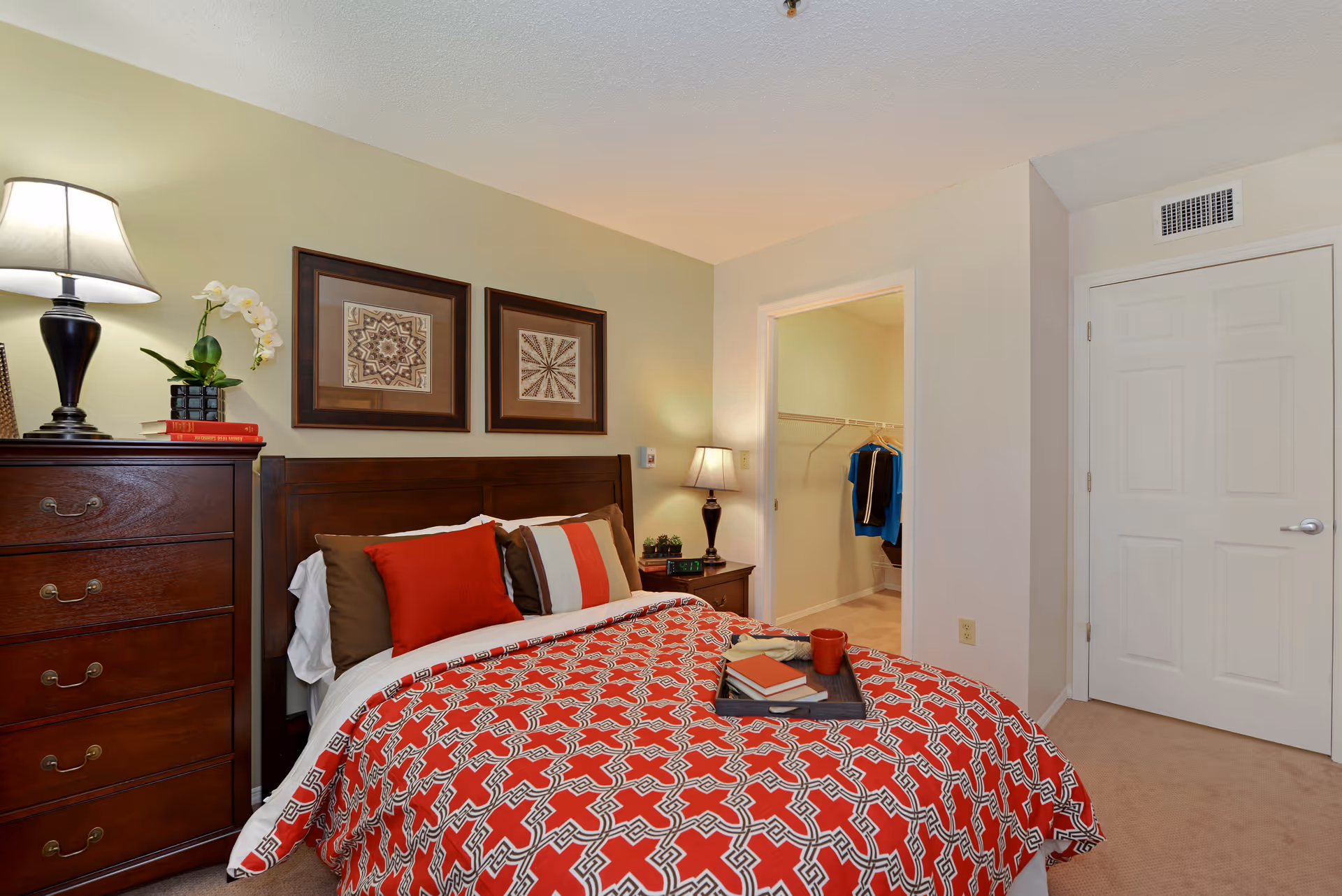 A cozy bedroom with a wooden bed frame and a red patterned bedspread. The bed has multiple pillows in red, brown, and white colors. On the left side, there is a wooden dresser with a lamp, a potted orchid, and some books. On the right side, there is a nightstand with a lamp, a clock, and small plants. The room has beige walls with two framed artworks above the bed. An open door reveals a walk-in closet with clothes hanging inside.