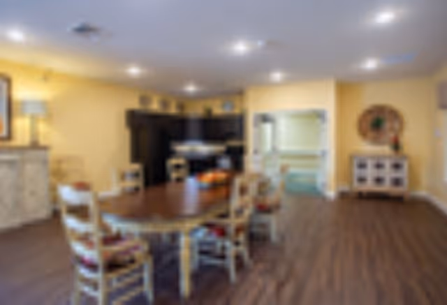 A spacious dining area with a large wooden table surrounded by six chairs with cushions. The room has yellow walls, wooden flooring, and recessed ceiling lights. In the background, there is a dark-colored kitchen area with cabinets and a countertop. A sideboard with a lamp and framed artwork is visible on the left, and a decorative mirror hangs on the right wall.