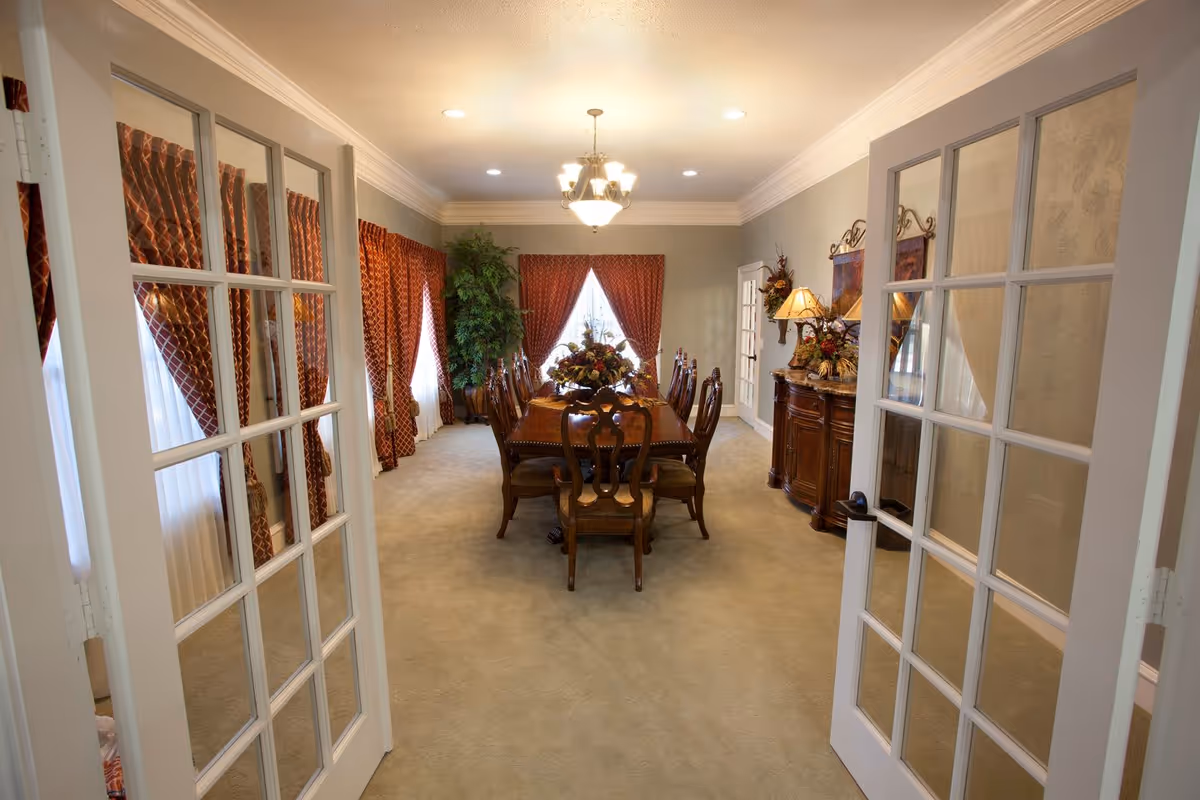 View through open double glass-paneled doors into an elegant dining room with a long wooden dining table surrounded by chairs, red patterned curtains on the windows, a chandelier overhead, and a sideboard with decorative items and a lamp.